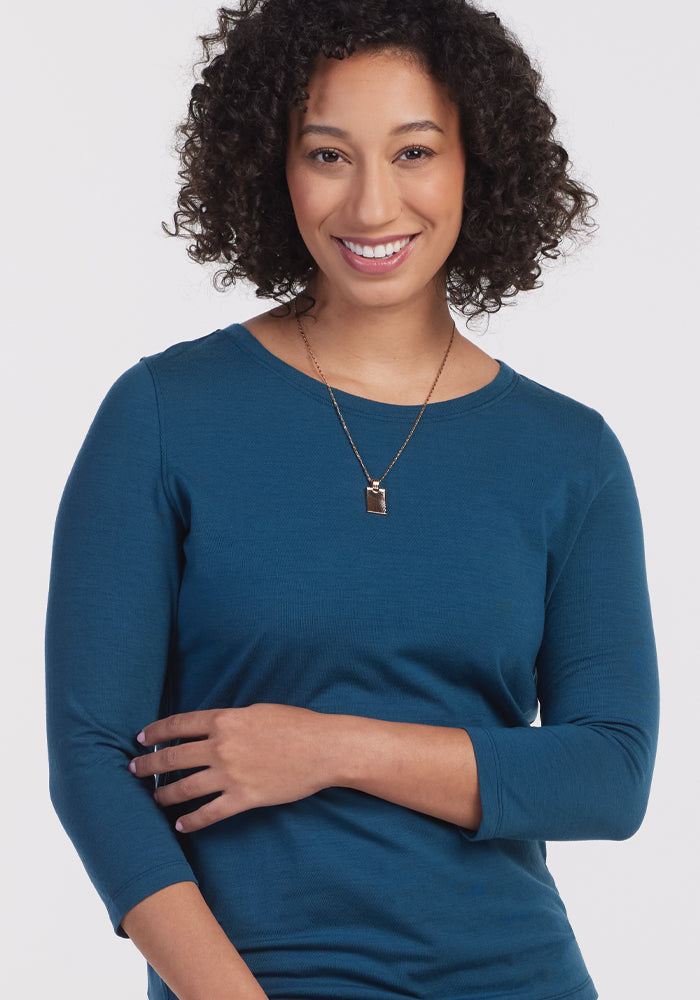 A person with curly hair smiles while wearing a Jenny 3/4 Sleeve Crew Neck in Real Teal from Woolx and a necklace with a pendant. The background is plain white.