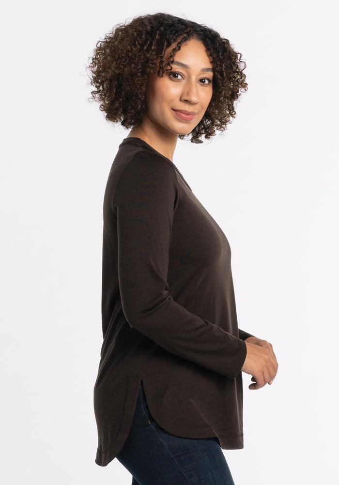 A person with curly hair stands sideways, wearing the lightweight Hazel Tunic in French Roast by Woolx, paired with dark jeans. The plain white backdrop enhances the elegant simplicity of their outfit.