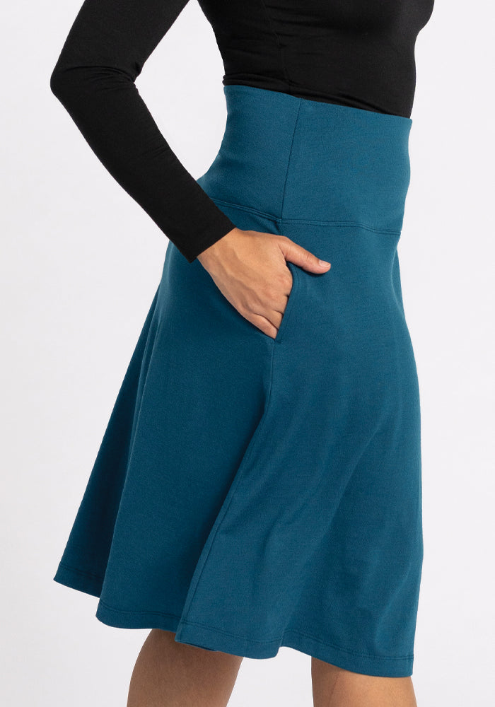 A person wearing the Marilyn Flowy Midi Skirt in Real Teal by Woolx, paired with a black long-sleeve top, stands against a plain background. The high-waisted skirt's merino wool design is visible from waist to knee and features convenient pockets.