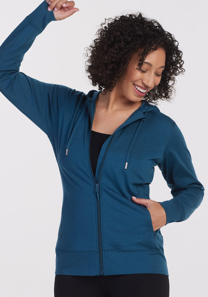 A person with curly hair is smiling and raising one arm while wearing the Ryann Hoodie in Real Teal from Woolx, layered over a black top. The athletic fit hoodie features drawstrings and pockets, set against a plain white background.