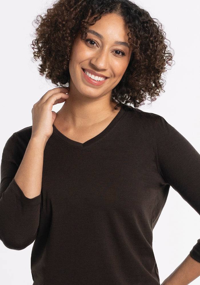 A person with curly hair is smiling and posing against a plain background. They are wearing a relaxed-fit, dark long-sleeved Elena V Neck in French Roast by Woolx and gently touching their neck with one hand.