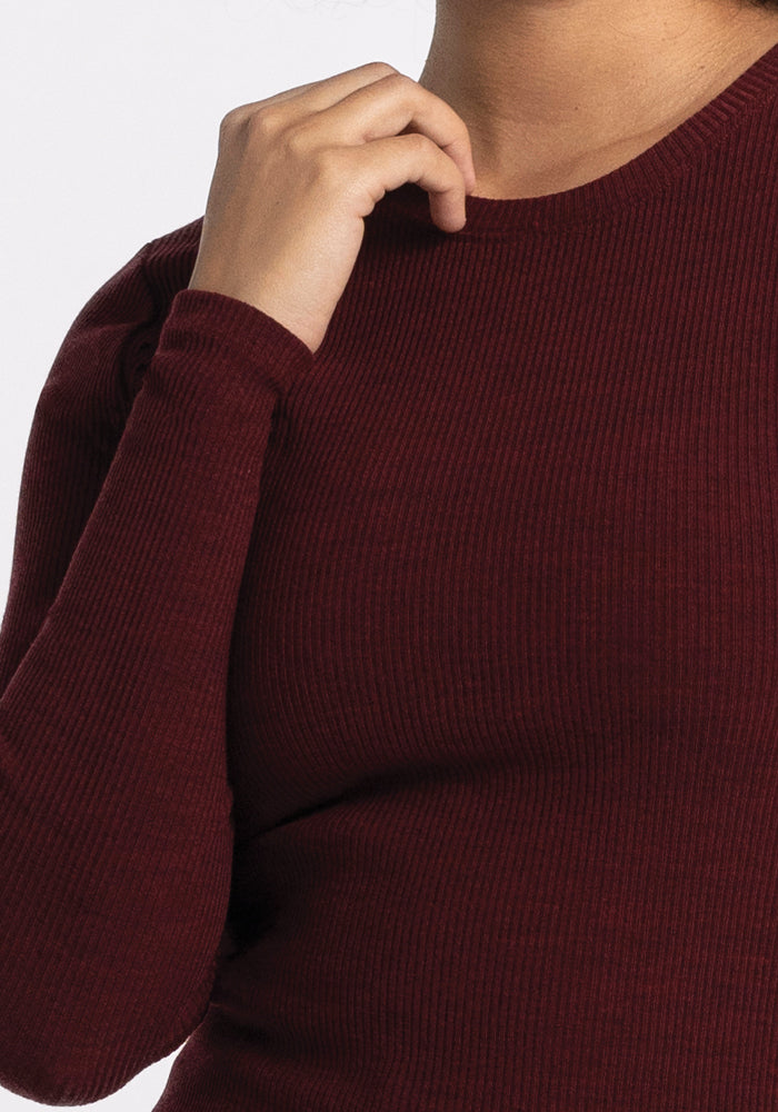 A person wearing the Hadley Ribbed Crew - Cranberry Melange by Woolx is depicted with their hand gently resting near their neck. Ideal for a travel wardrobe, the moisture-wicking material provides comfort against the plain white backdrop.