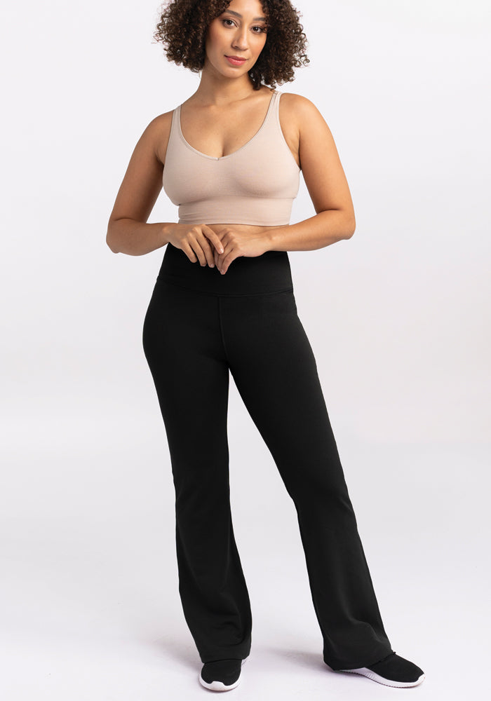 A person dressed in a beige tank top and Woolx's Marissa Pants in black stands confidently with hands clasped in front. They have curly hair and are sporting black and white sneakers, posing against a plain white background.