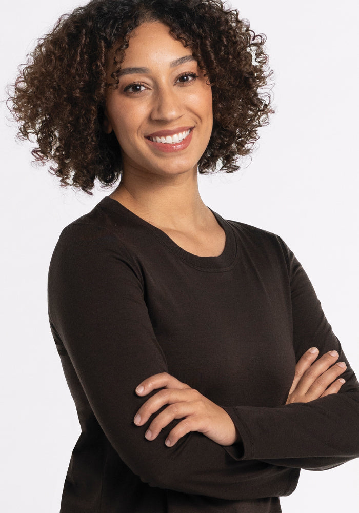 A smiling person with curly hair is wearing the Hazel Tunic in French Roast by Woolx, a black long-sleeve shirt made from lightweight merino wool, their arms crossed against a plain white background.