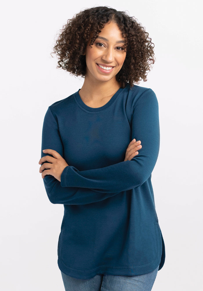 A person with curly hair smiles while crossing their arms. They are wearing the Woolx Hazel Tunic in Real Teal and jeans, standing against a plain white background.