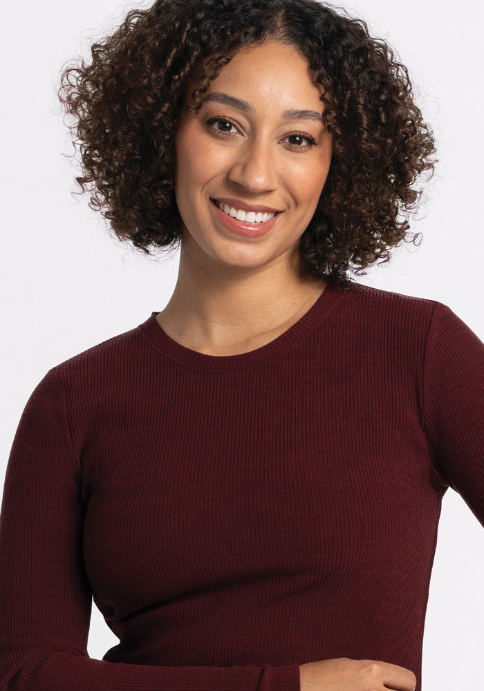 A person with curly hair wearing the Hadley Ribbed Crew in Cranberry Melange by Woolx, made from odor-resistant Merino wool, smiles at the camera against a plain white background, ready for any travel wardrobe.