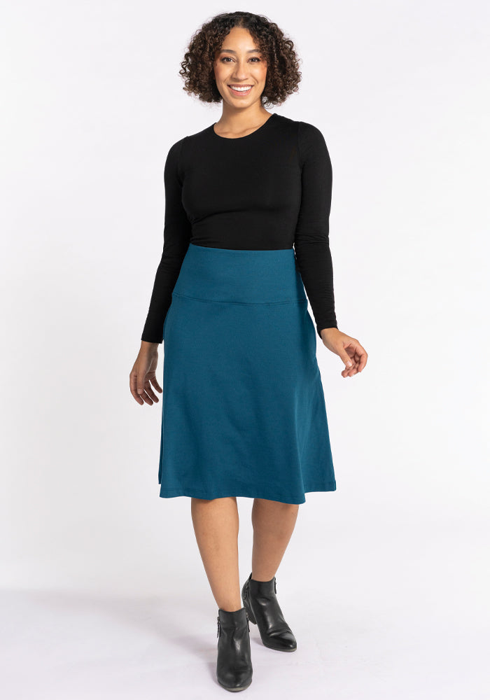 A person with curly hair smiles while wearing a black long-sleeve top and the Woolx Marilyn Flowy Midi Skirt in Real Teal. They are also wearing black ankle boots, standing against a plain white background.