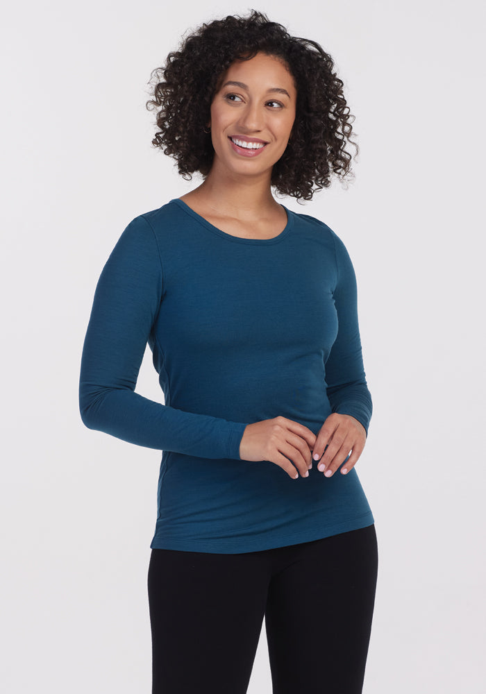 A person with curly hair wearing the Woolx Remi Long Sleeve T-Shirt in Real Teal, crafted from ultra-lightweight fabric, paired with black pants, stands smiling against a plain white background. 