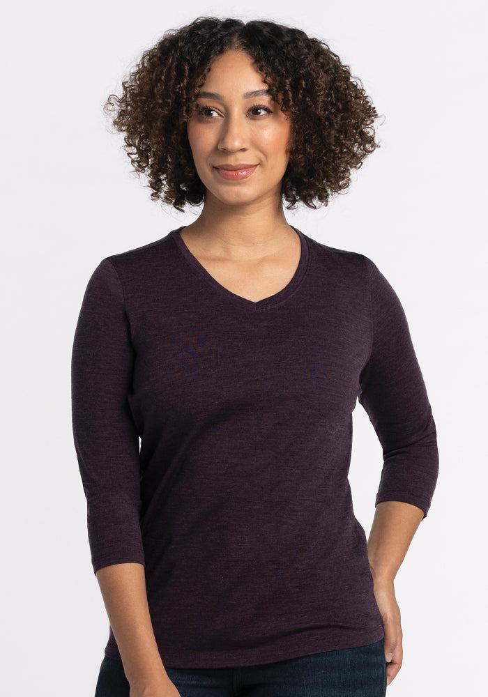 A person with curly hair is smiling and striking a pose with one hand on their hip. They are wearing a relaxed fit Elena V Neck in Deep Plum by Woolx, paired with jeans, against a plain light gray background.