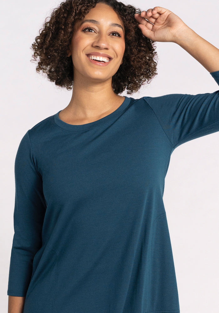 A person with curly hair is smiling and wearing the travel-ready Rissa A Line Dress in Real Teal from Woolx. Made from merino wool, the dress is perfect for any journey. They have one arm raised, touching their head against a plain background.