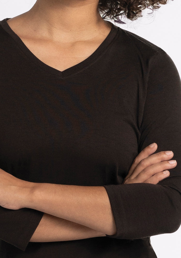 A person with curly hair is crossing their arms, wearing a French Roast Elena V Neck by Woolx in a relaxed fit. The background is a plain white.