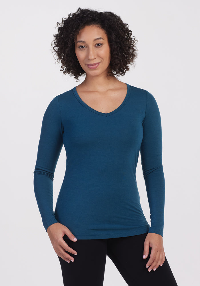 A person with curly hair is dressed in the fitted, long-sleeve Layla V Neck Top in Real Teal by Woolx and black pants, standing against a plain white background, smiling gently. 