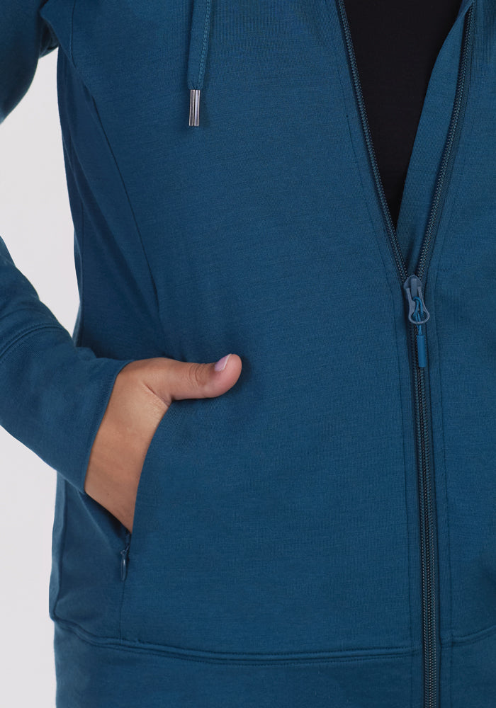 Close-up of a person wearing the Woolx Ryann Hoodie in Real Teal, featuring an athletic fit with their left hand in the pocket. The lightweight zip-up design made from Merino wool reveals a partially visible zipper, and the hood strings hang down against a plain white background.