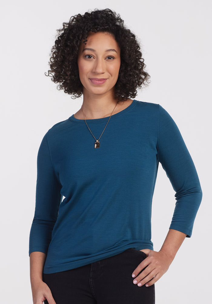 Smiling person with curly hair wearing a Jenny 3/4 Sleeve Crew Neck in Real Teal by Woolx and a necklace, standing against a plain white background.