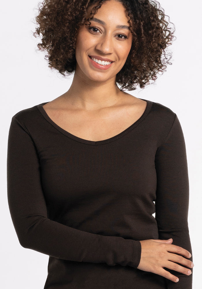 A woman with curly hair wearing the Layla V Neck Top in French Roast by Woolx smiles at the camera against a plain background. The breezy knit top drapes effortlessly, and her arms are relaxed at her sides.