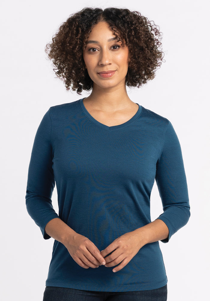 A person with curly hair is wearing a Real Teal Elena V Neck by Woolx, known for its relaxed fit. They are standing against a plain white background. 