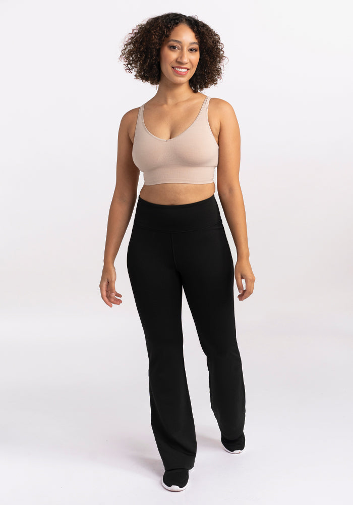 A person with curly hair smiles and walks forward, wearing a beige sports bra and the Marissa Pants in black from Woolx. The background is plain white.