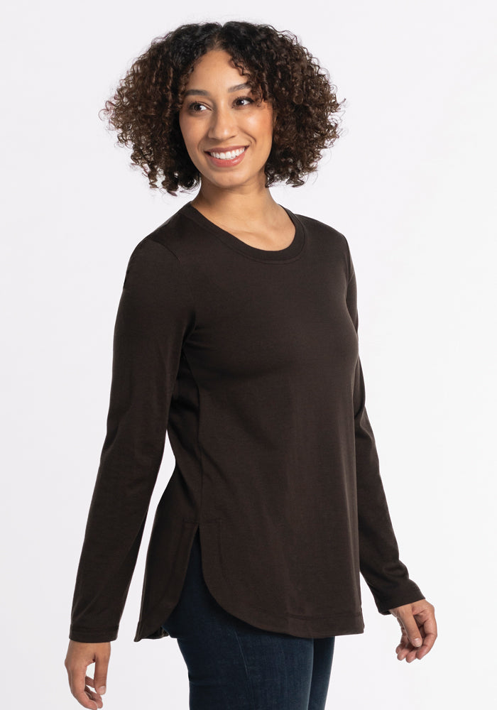 A person with curly hair smiles while wearing the Woolx Hazel Tunic in French Roast, a long-sleeved top made of lightweight fabric. The tunic features a round neckline and side slits as they stand against a plain white background. 