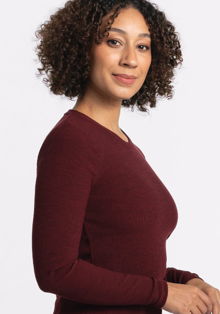 A person with curly hair is smiling at the camera, wearing a Woolx Hadley Ribbed Crew in Cranberry Melange, which makes a perfect addition to any travel wardrobe. The background is plain white.