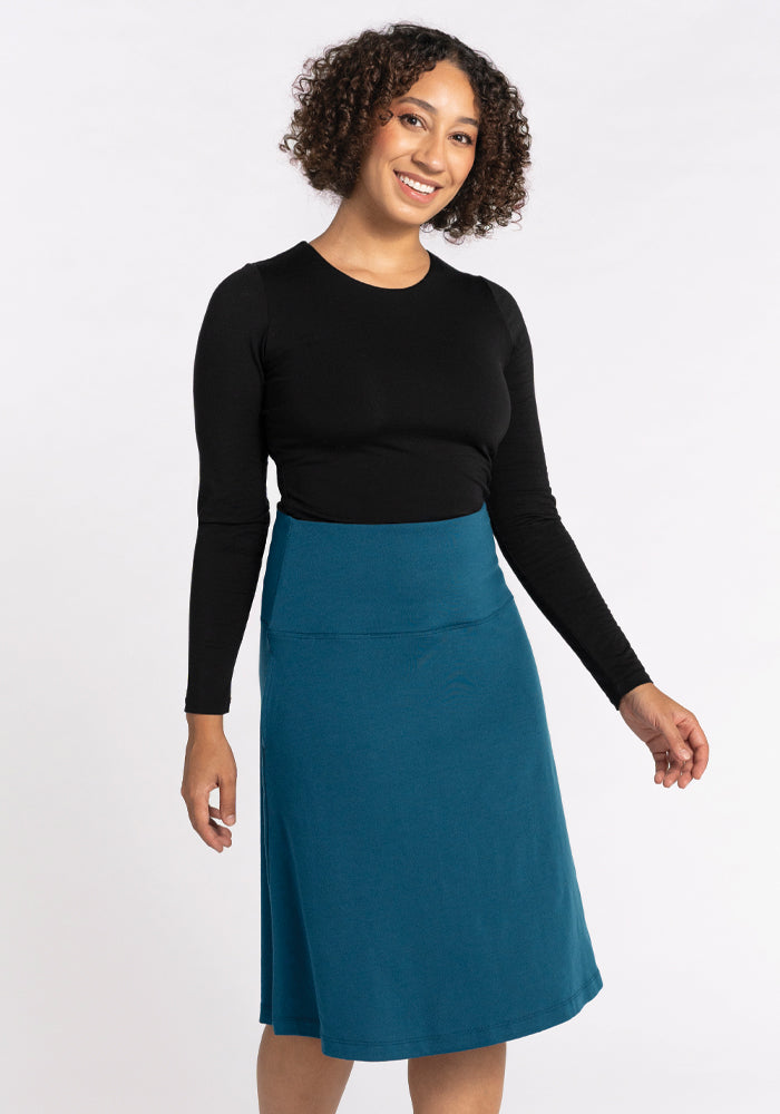 A person with curly hair smiles at the camera, donning a black long-sleeve top and the Marilyn Flowy Midi Skirt in real teal by Woolx. The background is plain white.