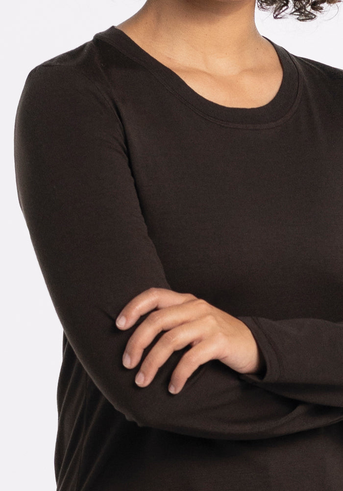 A person with curly hair is shown from the neck down, wearing the long-sleeved Hazel Tunic - French Roast by Woolx, crafted from lightweight fabric. Their arms are crossed against a plain white background.