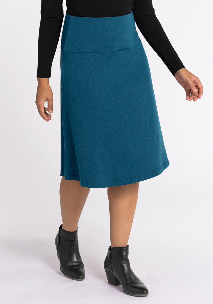 A person wearing a Woolx Marilyn Flowy Midi Skirt in Real Teal, paired with a black long-sleeve top and black ankle boots. The A-Line design of the outfit stands out against a plain white background. 