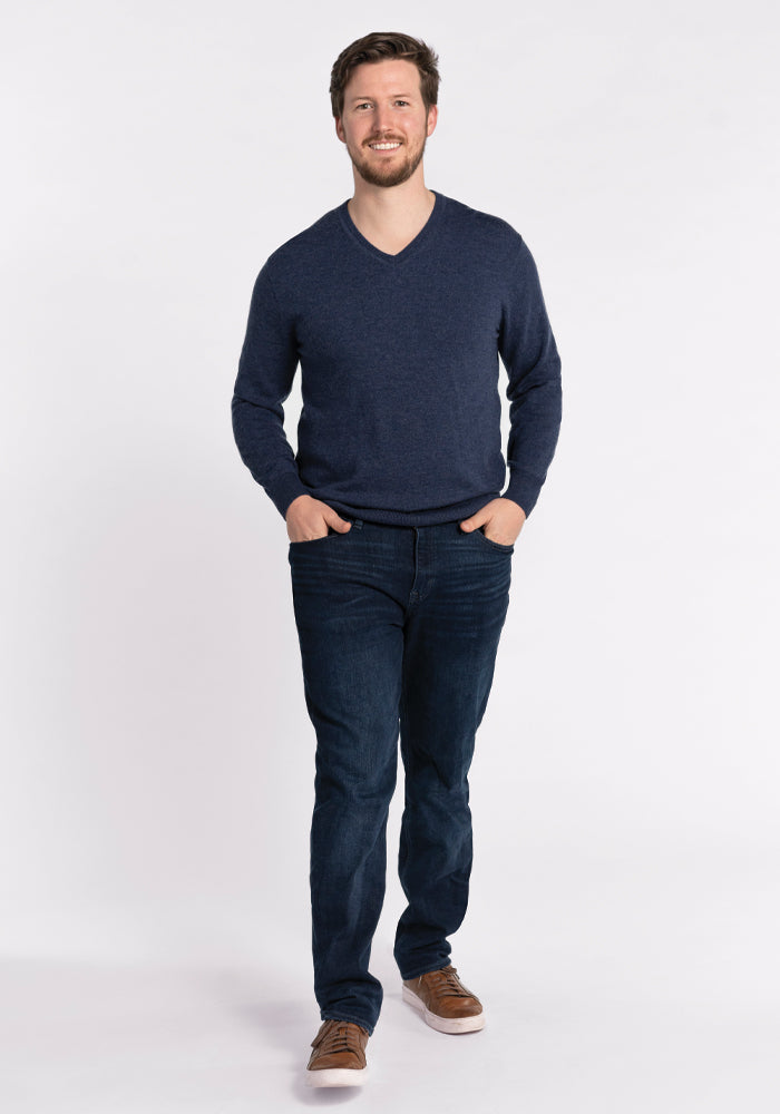 A man wearing the Vincenzo Sweater in Twilight Storm by Woolx, paired with dark jeans and brown shoes, stands smiling with his hands in his pockets against a plain white background.