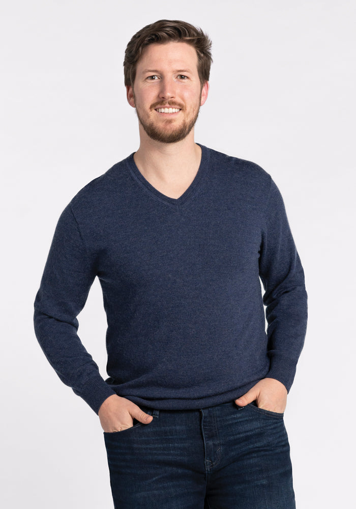 A man with a beard and short hair smiles at the camera, wearing the Vincenzo Sweater in Twilight Storm by Woolx along with dark jeans, standing against a plain white background with his hands in his pockets. 