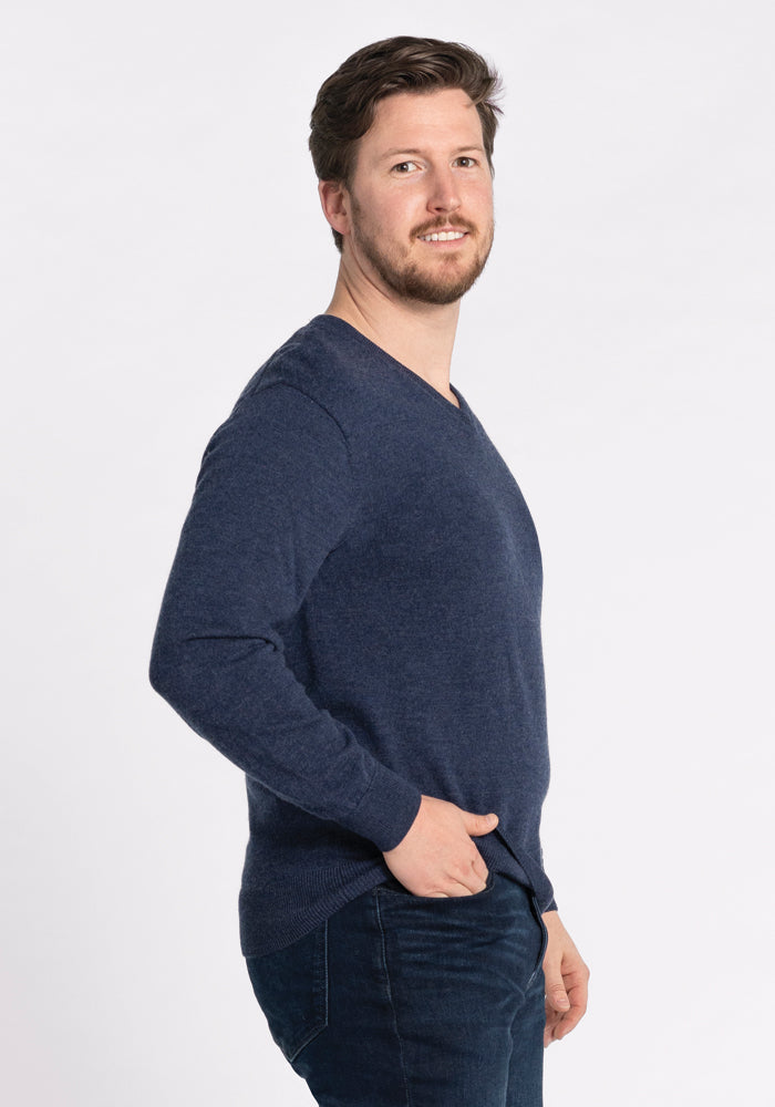 A man with a beard and short hair, dressed in the Woolx Vincenzo Sweater in Twilight Storm and jeans, stands sideways with one hand in his pocket, smiling at the camera against a plain white background.