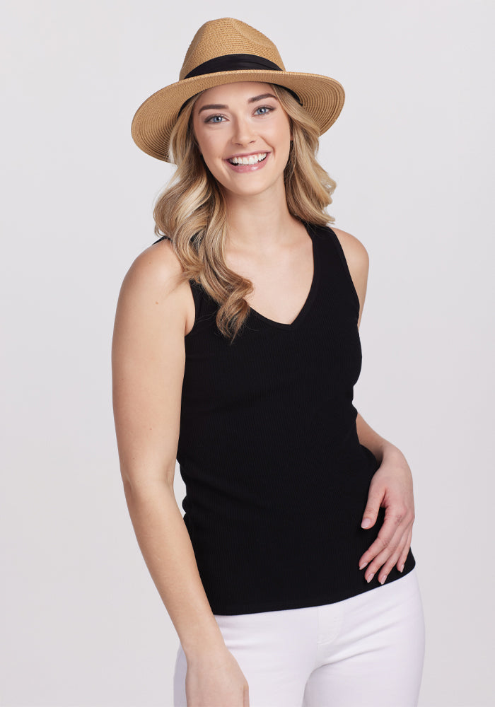 A woman in a Black Vivi Ribbed Tank by Woolx and a straw hat smiles at the camera, with long, wavy hair that captures capsule wardrobe vibes, against a plain light backdrop.