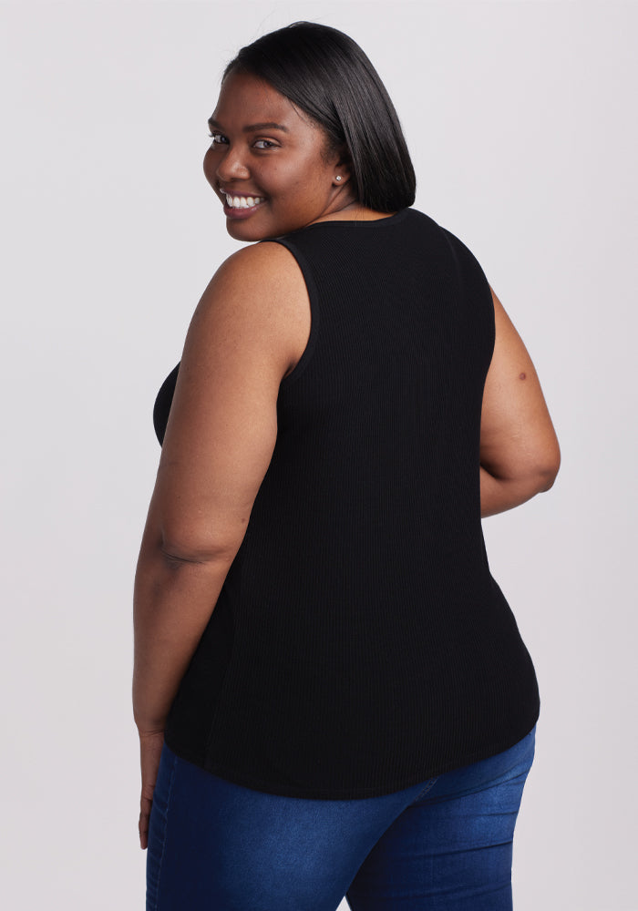 A woman in a sleeveless Vivi Ribbed Tank - Black by Woolx and blue jeans, embodies timeless chic and capsule wardrobe style as she smiles over her shoulder against a plain white background.