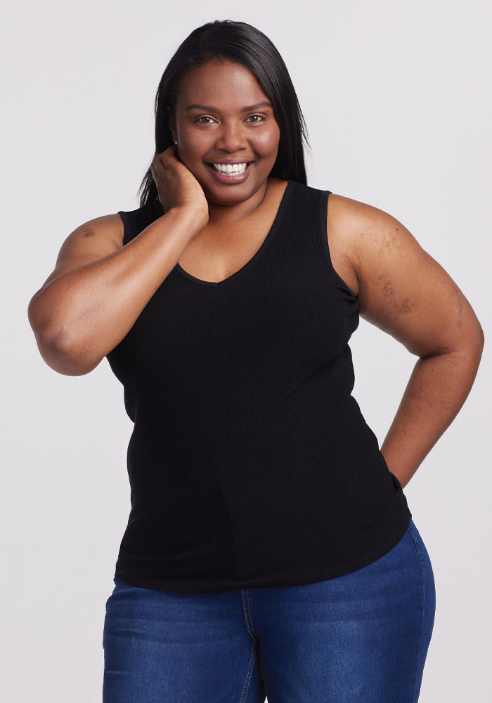 A person smiles, wearing a Woolx Vivi Ribbed Tank in black and blue jeans from their capsule wardrobe, with one hand on their hip and the other touching their face, set against a plain white background.