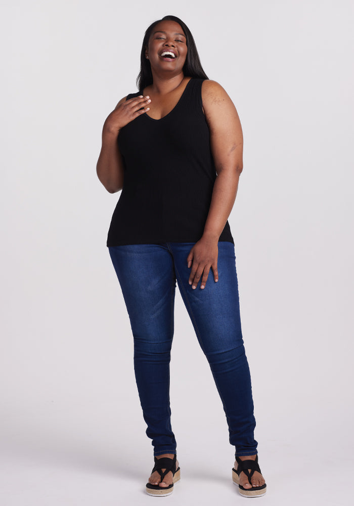 A woman with long hair laughs while wearing the Woolx Vivi Ribbed Tank in black, paired with blue jeans and wedge sandals. Her hand rests on her chest as she gazes upward, her capsule wardrobe style standing out against a plain white background.