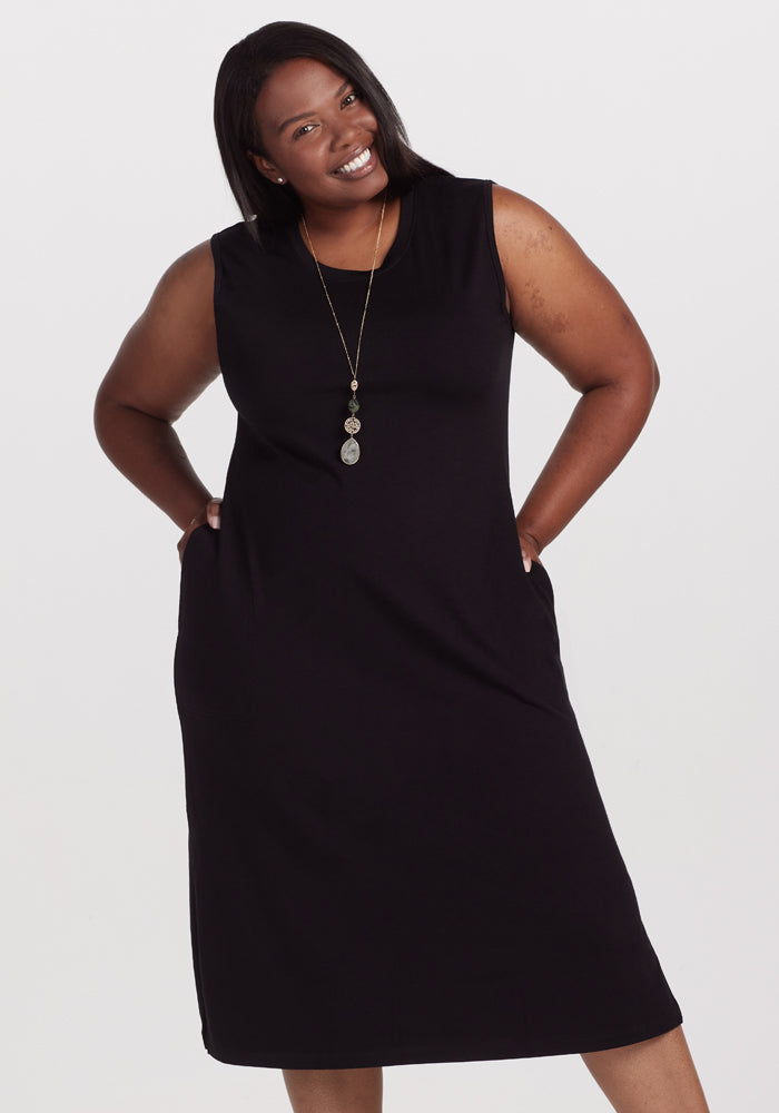 Against a plain white backdrop, a smiling woman confidently poses with hands on her hips, wearing the Woolx Cassie Dress in black. She accessorizes with a long necklace featuring three circular pendants. 