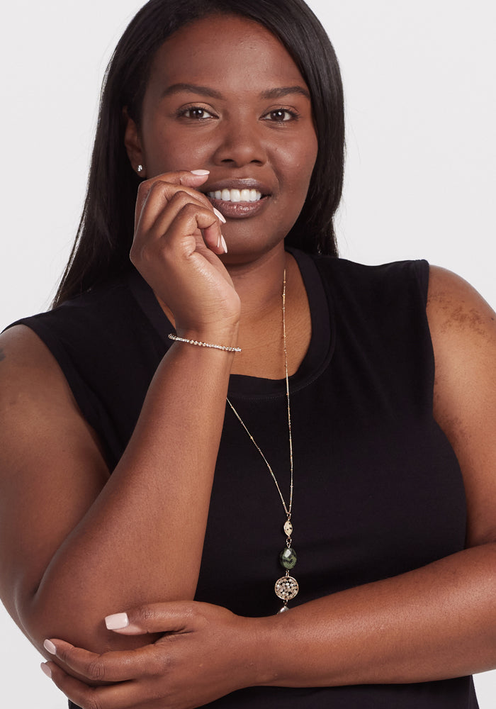 A woman with long dark hair smiles in the Cassie Dress - Black by Woolx. Sporting a long necklace with ornamental pendants and a bracelet, she gracefully poses with her right hand near her chin against a plain white background.