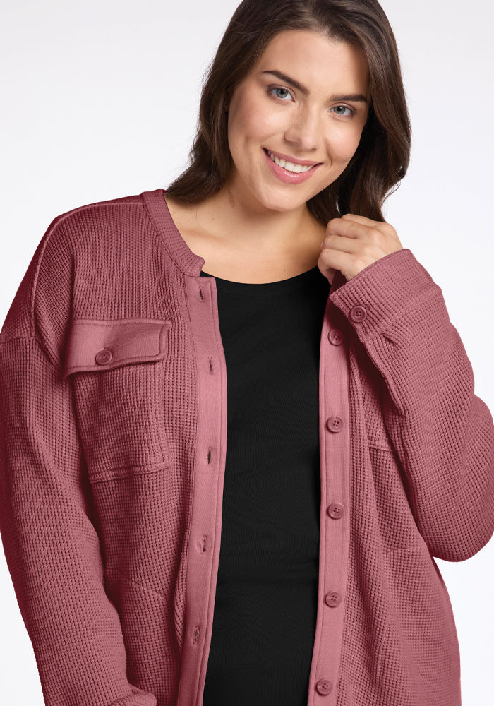 A person is smiling as they don a Wild Ginger Oaklynn Waffle Shacket from Woolx over a black top. Their long brown hair cascades gently as they hold the edge of this cozy piece. The plain white background accentuates the warmth of their attire.