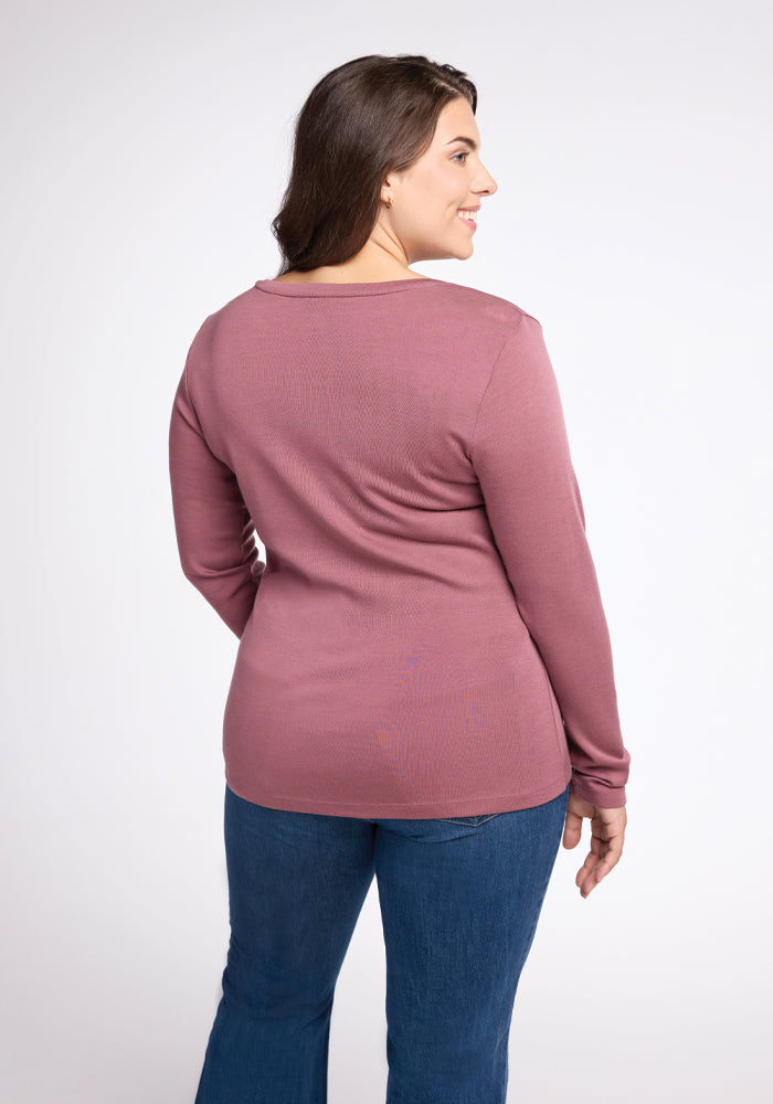 A woman with long, dark hair is standing with her back to the camera, smiling and looking to the side. She is wearing a Kenzie Scoop Neck in Wild Ginger from Woolx, paired with blue jeans, set against a plain light gray background.