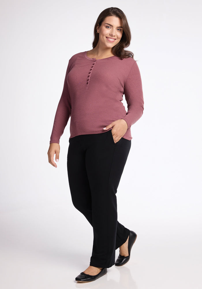 A woman in a Wild Ginger Reese Ribbed Henley by Woolx and black pants poses confidently in a studio setting. She stands with one hand in her pocket, wearing black flats, and smiles softly against a plain white background.