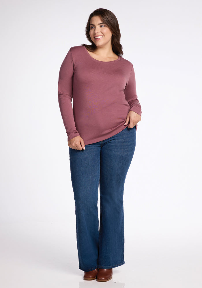 A person stands wearing the Kenzie Scoop Neck - Wild Ginger top from Woolx, paired with blue flared jeans and brown shoes. They have a relaxed pose and a slight smile against a plain white background.