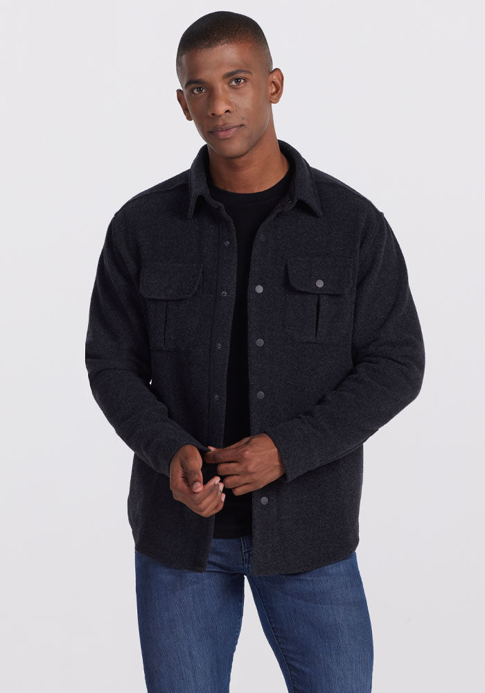 A man wearing the Woolx Wilder Shirt Jac in Carbon Black over a black t-shirt and blue jeans stands against a plain white background, exuding ultra-functional style as he looks directly at the camera. 