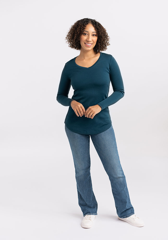 A person with curly hair is standing and smiling. They are wearing a long-sleeved, dark teal Merino wool Eva Tunic from Woolx, blue jeans, and white sneakers—perfect for a relaxed street style look. The background is plain and light-colored.