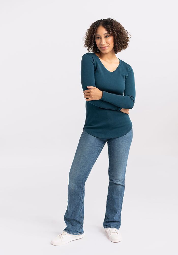 A person with curly hair stands with crossed arms, wearing a teal long-sleeve Eva Tunic from Woolx, blue jeans, and white sneakers. They are smiling slightly, embodying effortless street style against a plain light gray background.