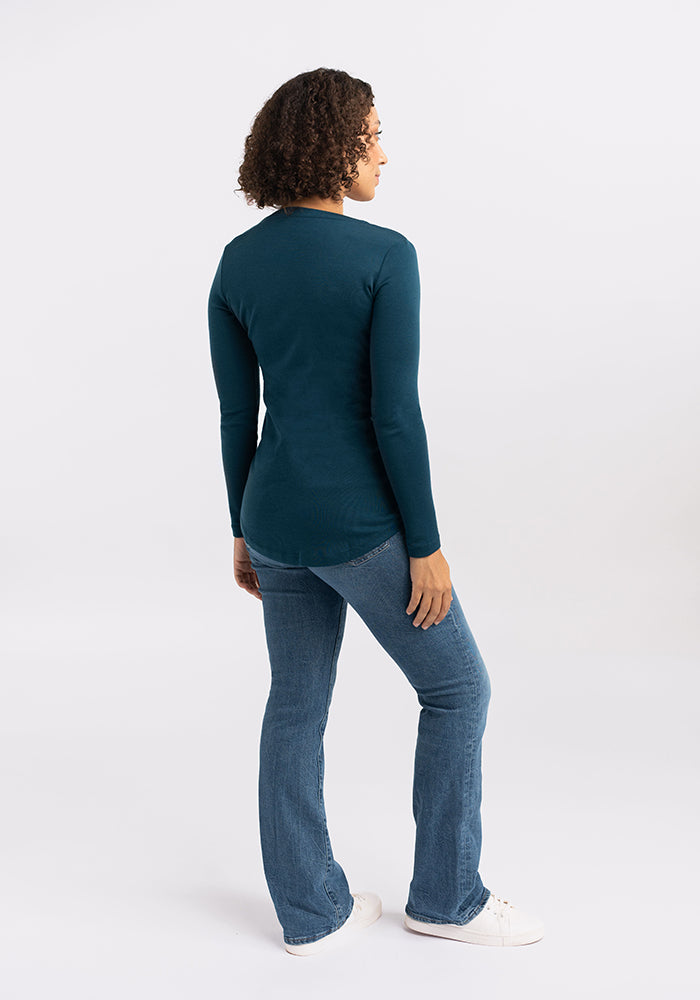 A person with curly hair stands facing away from the camera. They are wearing a long-sleeved, dark teal Eva Tunic by Woolx, blue jeans, and white sneakers. The background is plain and light-colored, showcasing a subtle street style vibe.