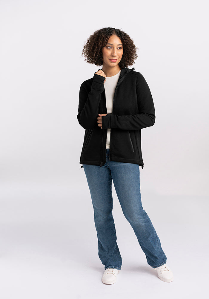 A person with curly hair stands against a white background, wearing a Woolx Cubby Hooded Sweatshirt in Black with heavy-duty zippers, a white shirt, blue jeans, and white sneakers. They are looking slightly to the side with their right hand partially covering their mouth.