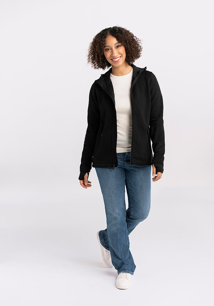 A person with curly hair is standing and smiling, wearing a Woolx Cubby Hooded Sweatshirt in Black, a white shirt, blue jeans, and white sneakers. They are posing with one leg slightly bent and a casual, relaxed expression. The background is plain white.