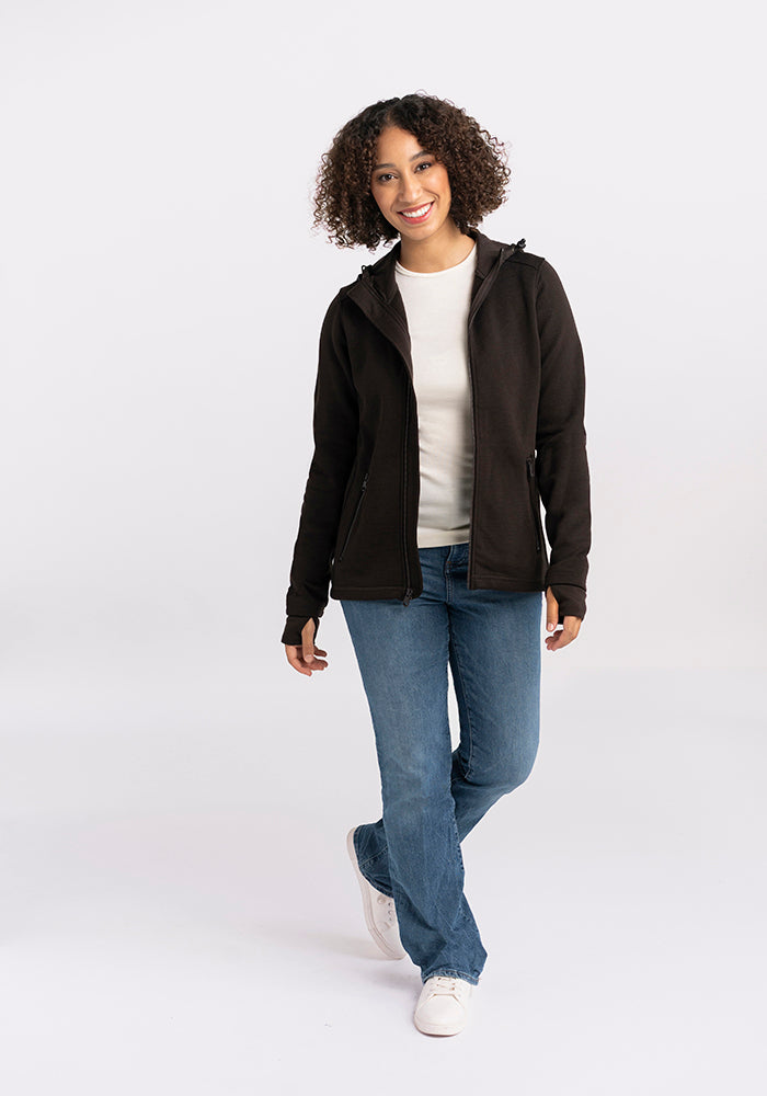 A person with curly hair is standing and smiling, wearing a black zip-up jacket over a white shirt, blue jeans, and white shoes. The coziest Cubby Hooded Sweatshirt - French Roast from Woolx made of Australian Merino Wool peeks out from underneath. The background is plain and light-colored.