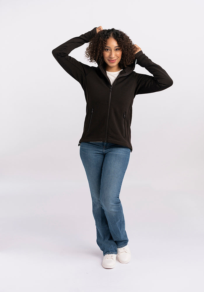 A person with curly hair stands against a plain background wearing a cozy Woolx Cubby Hooded Sweatshirt in French Roast, blue jeans, and white sneakers. They have their arms raised, adjusting the hood on their head, and they are smiling at the camera.