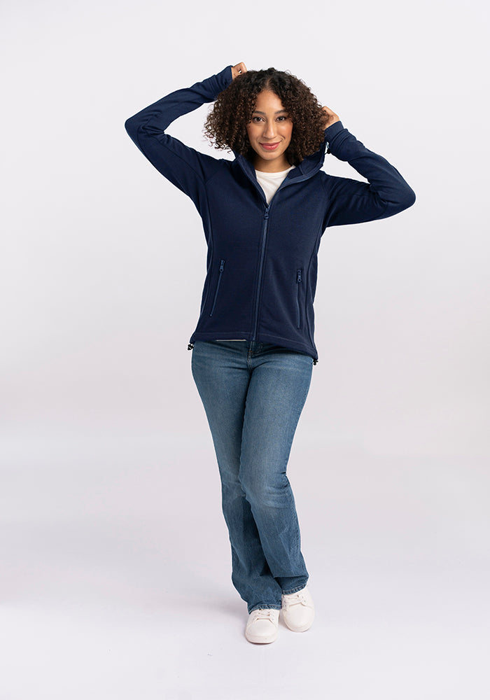 A person with curly hair, dressed in a cozy Woolx Cubby Hooded Sweatshirt in Deep Navy, light blue jeans, and white sneakers, stands against a plain white background. They have their hands raised, adjusting the hoodie while smiling warmly at the camera.