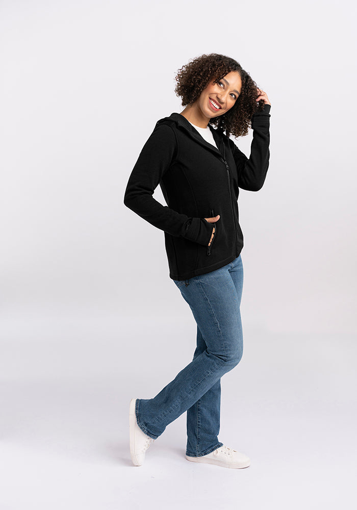 A person with curly hair is posing and smiling against a white background. They are wearing a Woolx Cubby Hooded Sweatshirt in Black with heavy-duty zippers, blue jeans, and white sneakers. One hand is touching their hair while the other rests in a pocket.