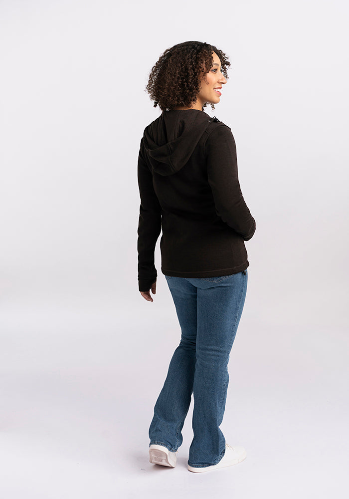 A person with curly hair, wearing a cozy Woolx Cubby Hooded Sweatshirt in French Roast, blue jeans, and white shoes is standing and facing slightly away from the camera. The background is plain white.